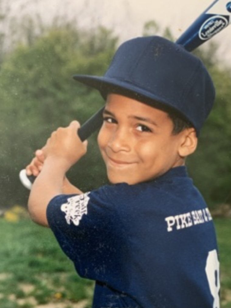 Micah Johnson as a young boy in a navy baseball cap, holding a bat over his shoulder and smiling.