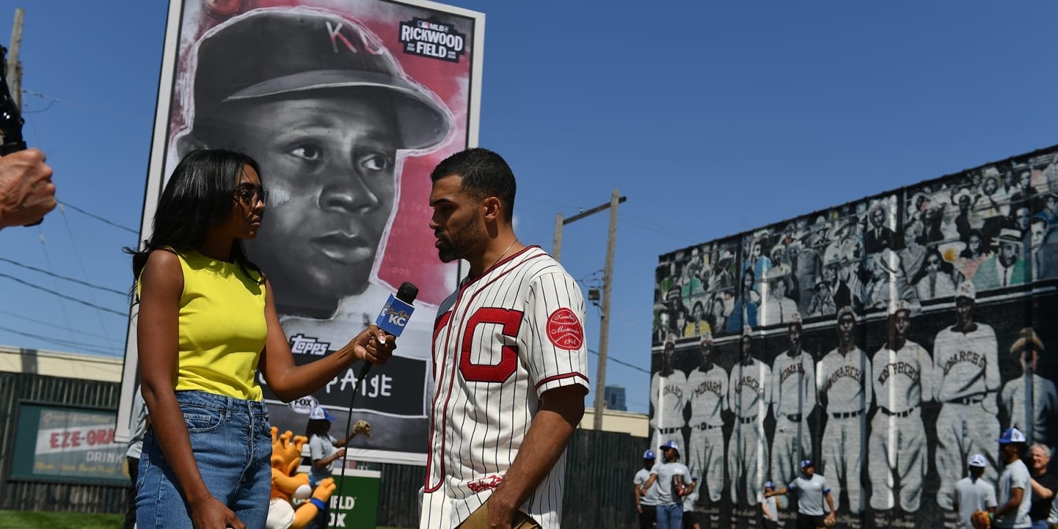 Micah Johnson, in a vintage baseball jersey, is interviewed in front of a large Satchel Paige baseball card bearing his artwork, with a mural of ballplayers behind them.