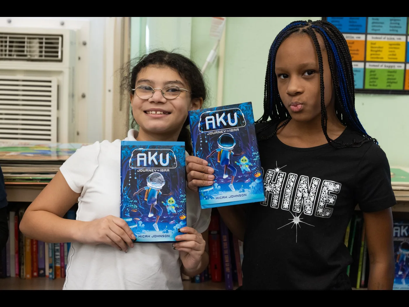 Two students hold up copies of Aku: Journey to Ibra in their classroom.
